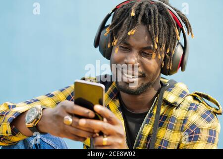 portrait of a young black man with a headphones writing on his smart phone with a blue wall in the background, technology and lifestyle concept, copy space for text Stock Photo