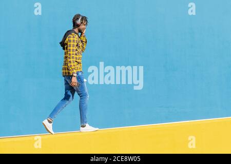 young black man with headphones walking along a yellow wall and a blue wall in the background with his smartphone in his hand, technology and lifestyle concept, copy space for text Stock Photo