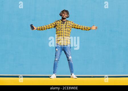attractive young black man listening to music from his phone with a large headphone on a blue background, technology and lifestyle concept, copy space for text Stock Photo