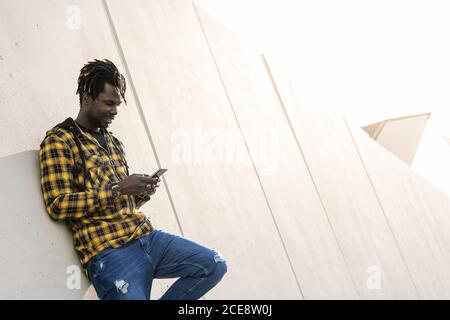 smiling young black man writing a message on his phone leaning on a modern concrete structure, concept of lifestyle and technology Stock Photo
