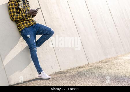 unrecognizable young black man writing a message on his phone leaning on a modern concrete structure, concept of lifestyle and technology Stock Photo