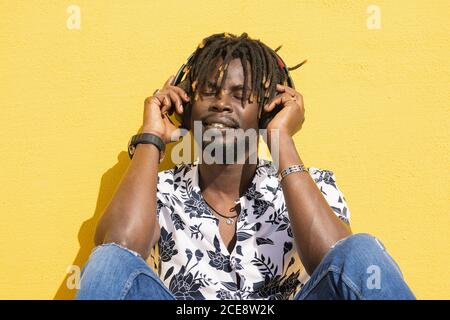 young black man sitting against a yellow wall listening and enjoying music with large headphones, technology and lifestyle concept Stock Photo