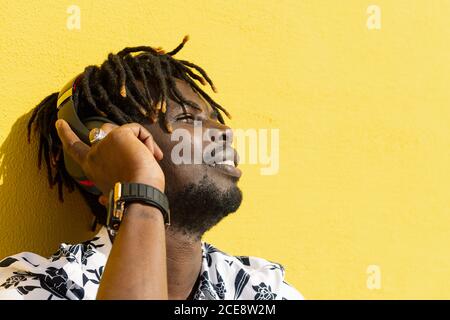 smiling young african man listening to and enjoying music with large headphones on yellow background, concept of technology and lifestyle, copy space for text Stock Photo