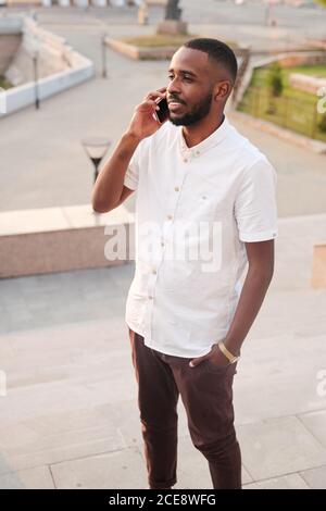 Content young Black manager in white shirt standing outdoors and holding hand in pocket while answering phone Stock Photo