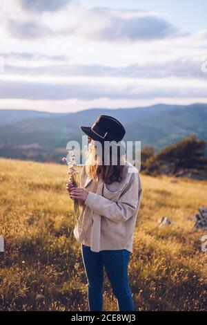 Tender female with bouquet of wildflowers standing on hill on background of majestic mountainous landscape in autumn during sundown Stock Photo