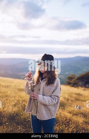 Tender female with bouquet of wildflowers standing on hill on background of majestic mountainous landscape in autumn during sundown Stock Photo