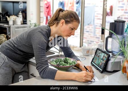 Woman in the kitchen with big bowl of salad watching zoom-meeting on electronic tablet and writes notes on paper Stock Photo