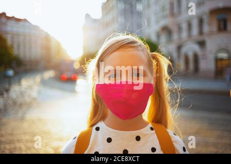 Life during covid-19 pandemic. Portrait of modern girl in white polka dot blouse with pink mask and yellow backpack going from school outdoors. Stock Photo