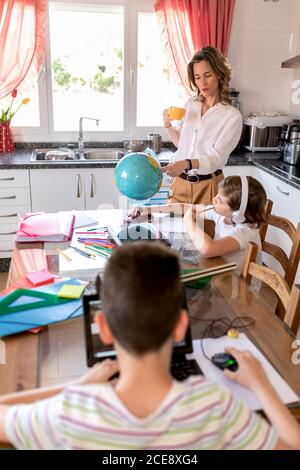 Adult female tutor pointing with finger at globe standing near concentrated pupil in headphones and near anonymous boy with laptop during geography lesson at home during geography lesson in kitchen Stock Photo
