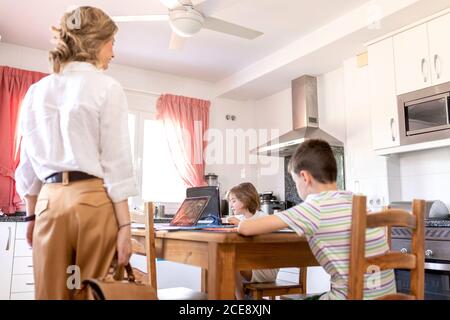 Back view of unrecognizable Woman in formal wear standing near siblings sitting at table while studying using netbook and tablet in modern style kitchen at home Stock Photo