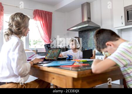 Concentrated mother in formal wear sitting leaned on hand while typing on netbook near unrecognizable son in headset and boy doing homework in light flat Stock Photo