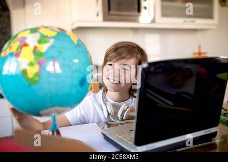 From above of cheerful child in casual wear sitting in front of netbook with headphones while looking at colorful shiny globe doing geography homework in apartment Stock Photo