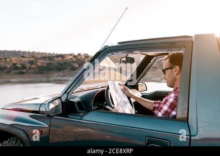 Side view of anonymous male traveler in casual wear and sunglasses watching map while sitting inside auto in front of river and mountains under calm sky Stock Photo