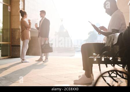 Busy black man using digital tablet and sitting on bench while colleagues drinking coffee in background Stock Photo