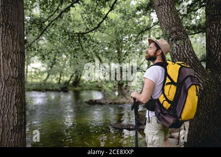 young man doing a hiking trail with his yellow backpack and hat on his head by a lake with many trees and natural areas looking at the landscape Stock Photo