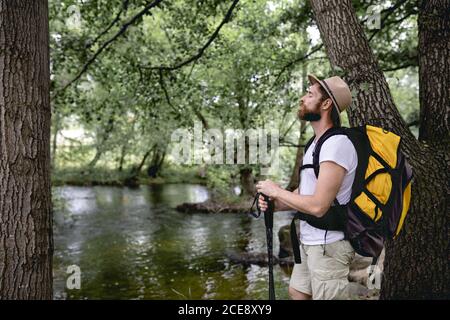 young man doing a hiking trail with his yellow backpack and hat on his head by a lake with many trees and natural areas looking at the landscape Stock Photo
