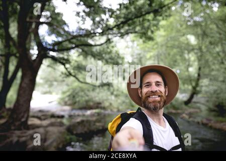 young man with beard hat on his head and yellow backpack hiking down a lake route with trees and shady areas taking a selfie Stock Photo
