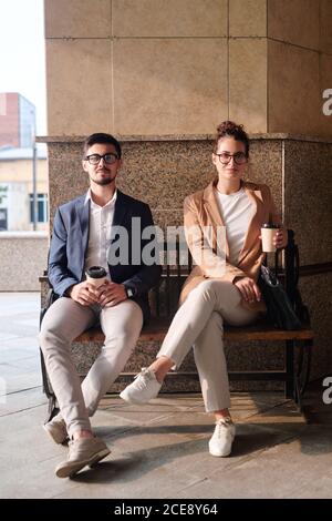 Portrait of content stylish business colleagues sitting on bench and drinking coffee from disposable cups Stock Photo