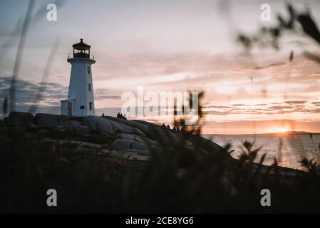 Silhouettes of anonymous people sitting on rocky coast near Peggys Cove Lighthouse and admiring cloudy sunset sky over sea in evening in Canada Stock Photo