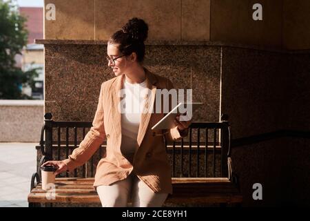 Smiling attractive young businesswoman sitting on bench and drinking coffee while working with tablet outdoors Stock Photo