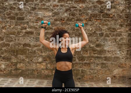 Woman athlete in sports clothes training shoulders using dumbbells near rough wall Stock Photo