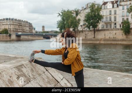Side view of fit ethnic female athlete listening to music in wireless headphones while stretching leg during workout on embankment near pond and building in town under cloudy sky Stock Photo