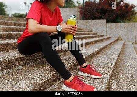 Side view of anonymous fit ethnic female athlete in sports clothes and smart watch drinking water from thermos while resting with closed eyes on staircase after training in sunlight
