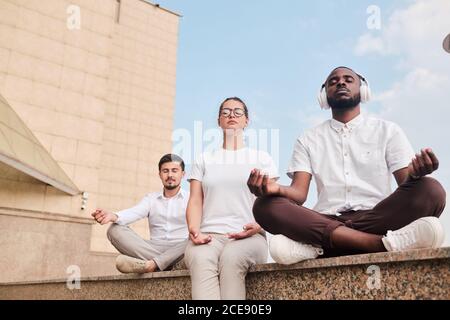 Serene young multi-ethnic people sitting on stone railing and meditating with closed eyes in city Stock Photo