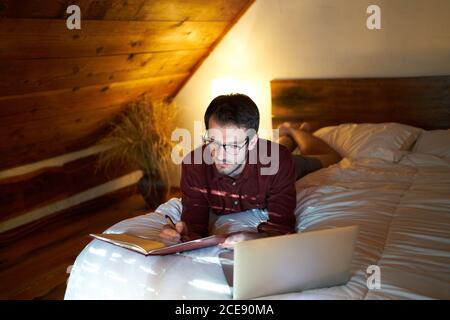Busy male freelancer lying on bed with laptop in cozy bedroom and writing in daily planner while working remotely at home Stock Photo