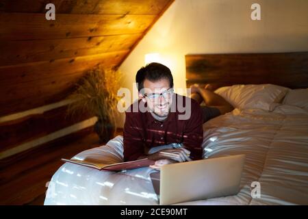 Busy male freelancer lying on bed with laptop in cozy bedroom and writing in daily planner while working remotely at home Stock Photo