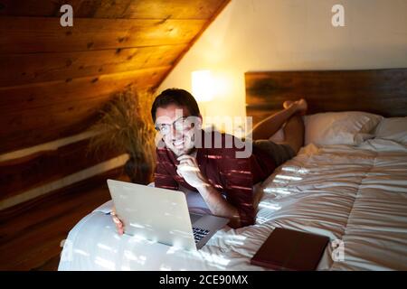 Thoughtful male entrepreneur in casual wear lying on bed and browsing netbook while working on remote project Stock Photo