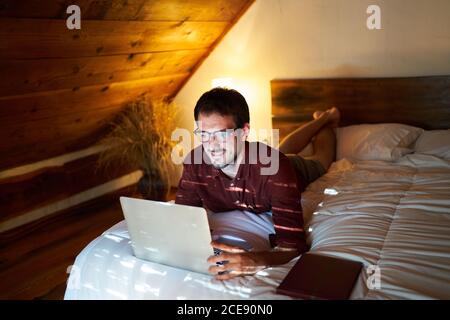 Thoughtful male entrepreneur in casual wear lying on bed and browsing netbook while working on remote project Stock Photo