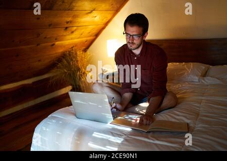 Busy male freelancer sitting on bed with laptop in cozy bedroom and writing in daily planner while working remotely at home Stock Photo