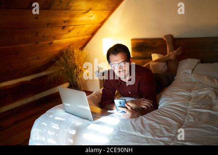 Thoughtful male entrepreneur in casual wear lying on bed and browsing netbook and using smartphone while working on remote project Stock Photo