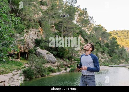 Concentrated male controlling modern drone flying on background of amazing mountainous landscape and lake Stock Photo