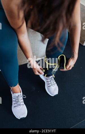 High angle of crop anonymous female athlete in sportswear adjusting elastic bandage on leg while preparing for training in gym
