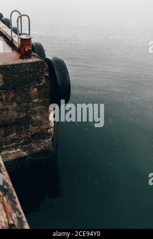 Crop part of stone pier with tires and chains in raindrops falling on water surface. Stock Photo