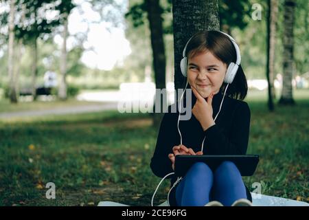 happy girl in white headphones using digital tablet pc in the park. Distant learning concept. Resilience, back to school, new normal. Stock Photo