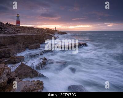 Waves crashing below a lighthouse at sunrise.