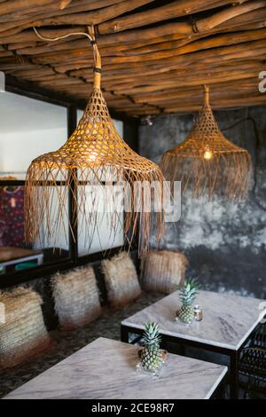 Trendy loft hipster cafe interior design. White marble tables, wicker straw lamps and pillows, bamboo roof, gray concrete walls. Stock Photo