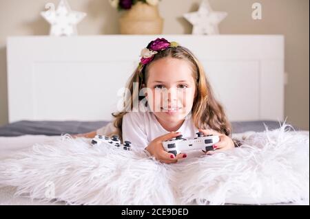 A little girl is playing a computer console on a sofa in a bright room. Concurrent games for children. Joystick for control in hands. Emotions and fun of the child. Copy space. High quality.Soft focus Stock Photo