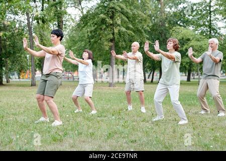 Focused coach raising hands forward while showing relaxing exercise to senior students at qigong practice Stock Photo