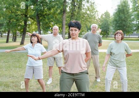 Mature brunette instructor moving arm aside and practicing qigong with senior students in park Stock Photo