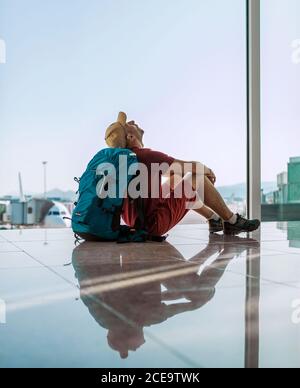 Alone backpacker traveler sitting on the airport terminal floor near the window, looking at the hall ceiling and waiting for boarding at aircraft whic Stock Photo