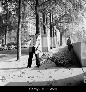 1950s, historical,  a man wearing civilian clothes and hat sweeping up fallen leaves lying on a pavement from the trees that line the avenues by the river Seine in Paris, France. Stock Photo