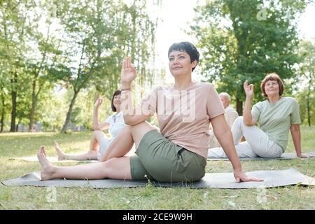 Attractive yoga coach with short hair sitting on mat and practicing marichis pose with students at group class Stock Photo