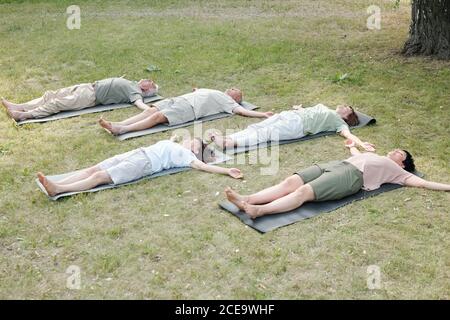 Group of mature yoga students lying in corpse pose on mats while resting after training in park Stock Photo