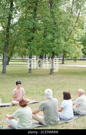 Middle-aged yoga instructor sitting on mat in park and explaining breathing technique to senior students Stock Photo