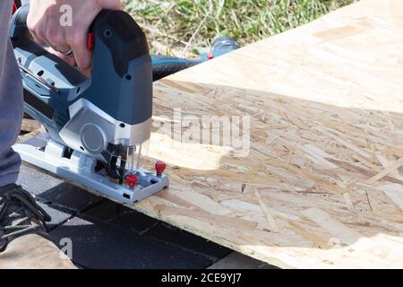 A young worker cuts a sheet of plywood with an electric jigsaw on the street. Stock Photo