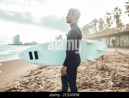 Side view of man wearing wetsuit and standing with surfing board on sandy beach, Spain Stock Photo
